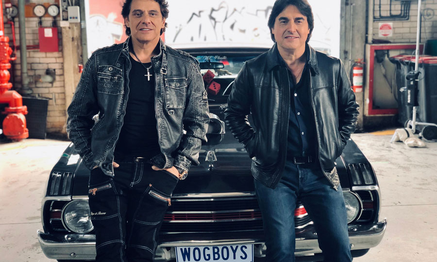 Two middle aged Greek Australian men sitting on the bonnet of a muscle car with the number plate WOGBOYS