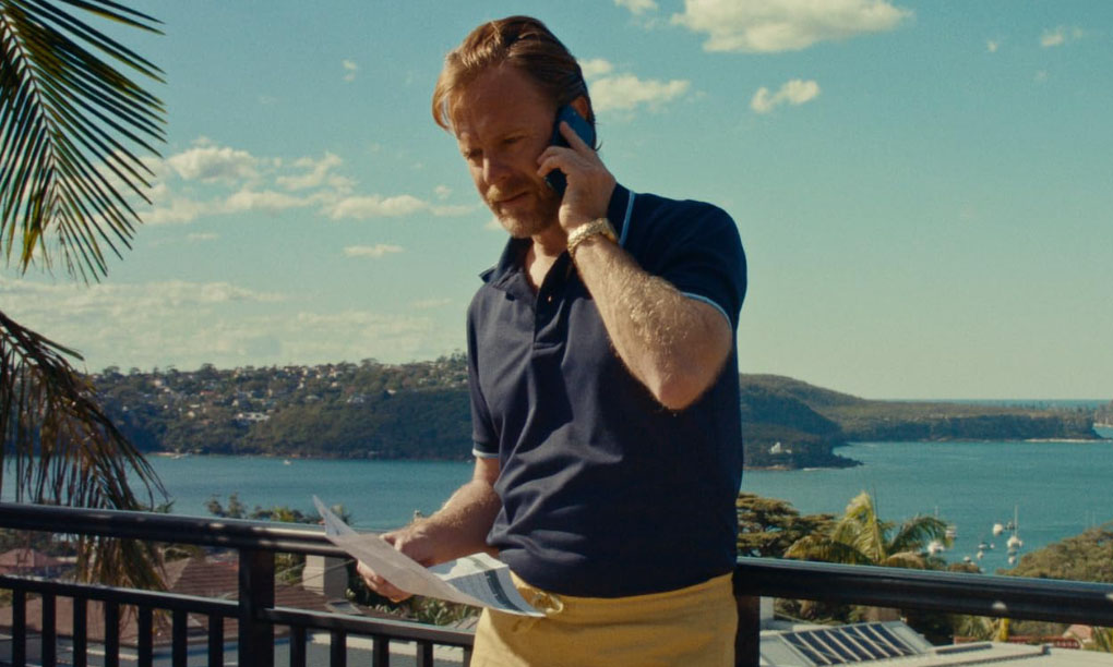 A man talking on a mobile phone standing a balcony with a harbour view behind him