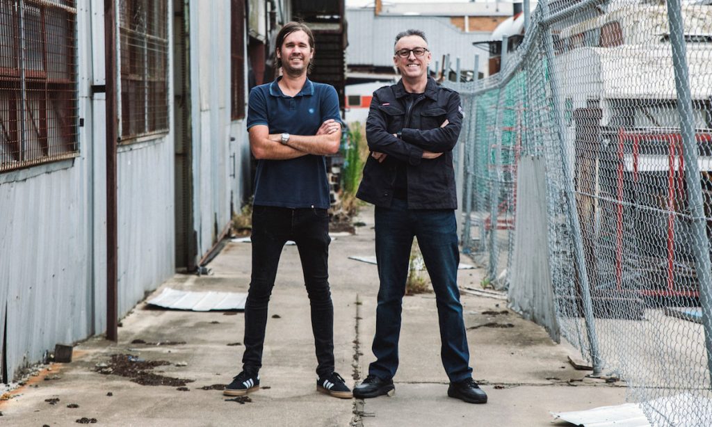Matt Dower and Tony Martin pose between a warehouse and a wire fence