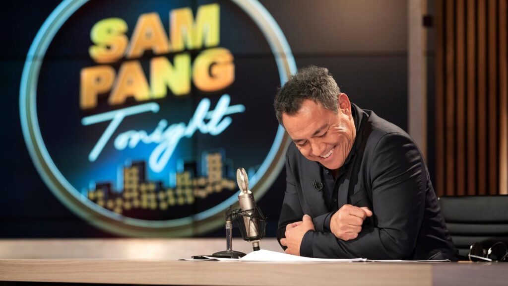 Sam Pang sitting at his tonight show desk laughing